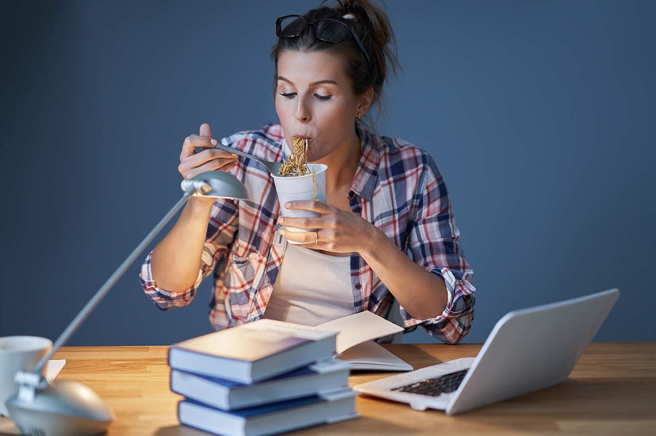 A student eating noodles at a desk stacked with books